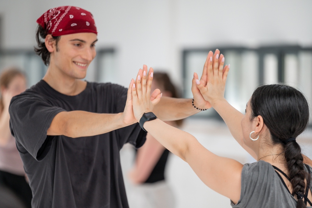 Two dancers face each other, touching palms out in front of them during a training session in a dance studio